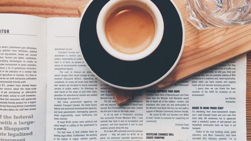 Eine Zeitung liegt auf einem Tisch, darauf steht eine Tassee Kaffee und ein Glas Wasser.